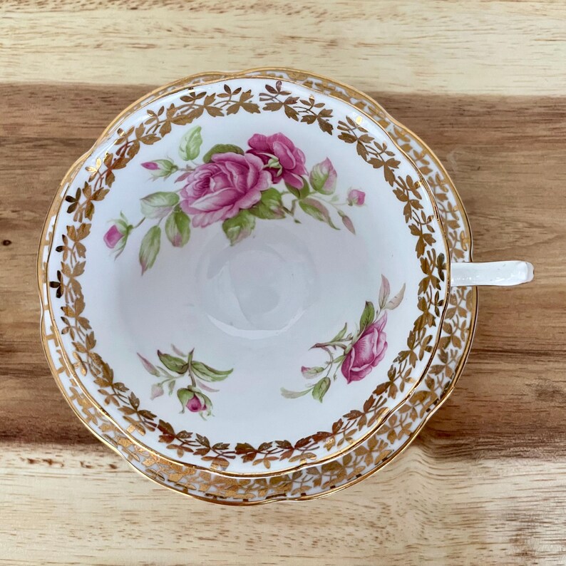 Puede incluir: Juego de taza y platillo blancos con dise&ntilde;o floral de rosas rosas. La taza y el platillo tienen un borde dorado y un asa blanca. El conjunto est&aacute; sobre una superficie de madera.