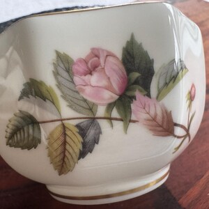 Pode incluir: Ch&aacute;vena de ch&aacute; hexagonal creme com uma borda dourada e um design floral. O design apresenta uma rosa rosa com folhas verdes e castanhas. A ch&aacute;vena de ch&aacute; &eacute; provavelmente feita de porcelana e &eacute; um item vintage.