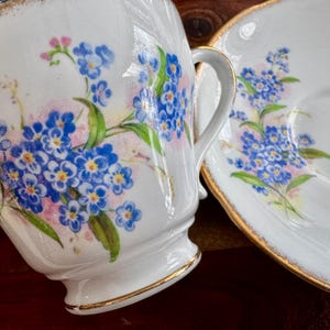 May include: A white teacup and saucer set with a floral design featuring blue forget-me-not flowers and green leaves. The set has a gold trim around the edges.