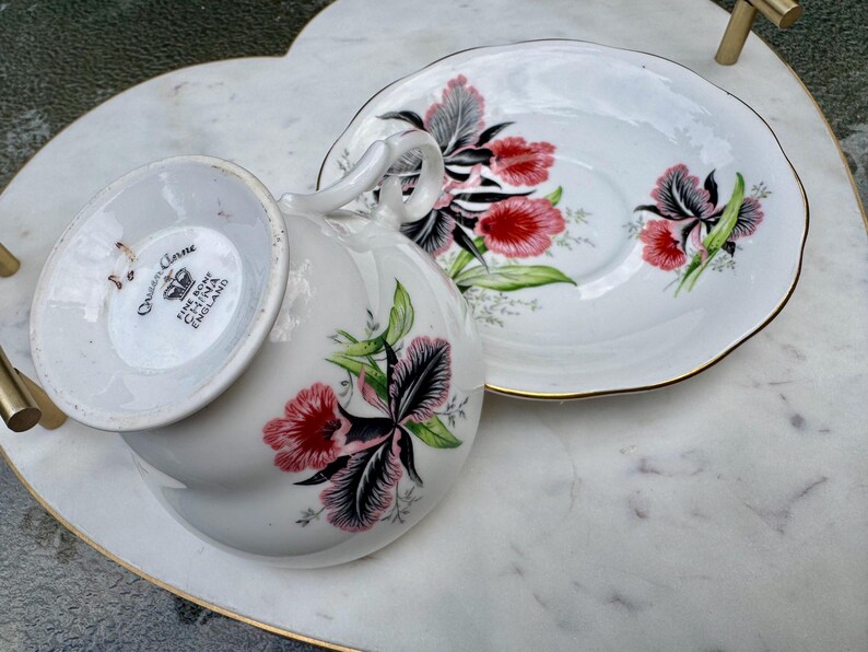 Puede incluir: Un juego de taza y platillo blancos con un dise&ntilde;o floral. La taza y el platillo est&aacute;n decorados con flores rojas, rosas y negras con hojas verdes. El juego tiene ribete dorado y se ven las palabras "Queen Anne Fine Bone China England".