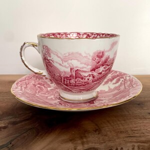 Pu&ograve; includere: Un set di tazza e piattino bianchi con un disegno di paesaggio rosso. La tazza e il piattino hanno finiture dorate. La tazza da t&egrave; &egrave; su una superficie di legno.