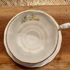 May include: A white teacup and saucer with a gold rim and a floral design featuring yellow flowers. The saucer has a scalloped edge.