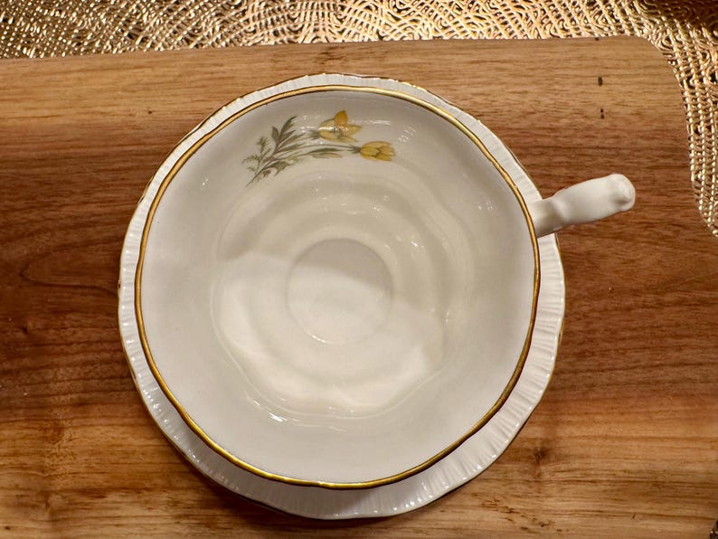 May include: A white teacup and saucer with a gold rim and a floral design featuring yellow flowers. The saucer has a scalloped edge.