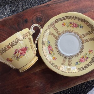 May include: A pale yellow porcelain teacup and matching saucer set featuring a delicate floral pattern with pink and white roses, accented by intricate gold leaf filigree borders and scalloped edges on both the cup and the saucer rim.