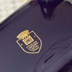 May include: Close-up of a dark blue porcelain object with a gold emblem. The emblem features a crown, building, and text that reads "Porcelaine Limoges Castel France". The object has a glossy finish.