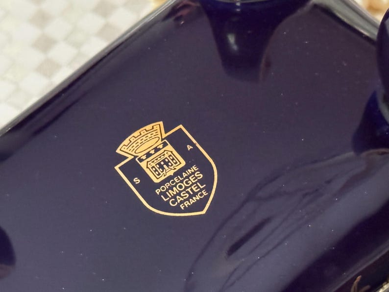 May include: Close-up of a dark blue porcelain object with a gold emblem. The emblem features a crown, building, and text that reads "Porcelaine Limoges Castel France". The object has a glossy finish.