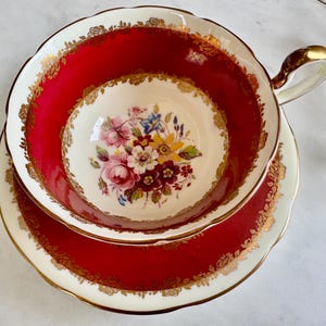 May include: A red teacup and saucer with a gold rim and floral pattern. The cup has a scalloped edge and a handle. The saucer has a matching design.