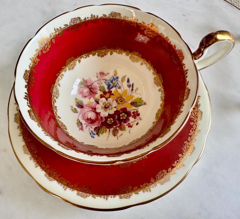 May include: A red teacup and saucer with a gold rim and floral pattern. The cup has a scalloped edge and a handle. The saucer has a matching design.