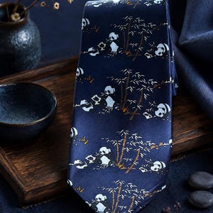 May include: A dark blue necktie featuring a repeating pattern of black and white pandas eating bamboo stalks. The design includes small brown accents near the base of the bamboo plants, set against a smooth, solid dark blue fabric background.