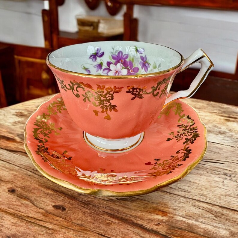 May include: A coral pink teacup and saucer with gold trim and a floral design. The teacup has a white interior with a floral pattern in shades of purple and green.