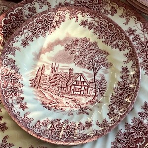 Pu&ograve; includere: Un set di piatti antichi con un design floreale e paesaggistico rosso e bianco. I piatti presentano una scena centrale di una casa e alberi, circondata da un bordo decorativo di foglie e uva. Un set di stoviglie classico ed elegante.
