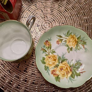 May include: A green and white teacup and saucer set with a floral design featuring yellow roses. The saucer has a scalloped edge.