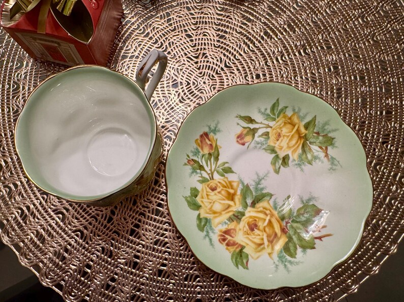 May include: A green and white teacup and saucer set with a floral design featuring yellow roses. The saucer has a scalloped edge.