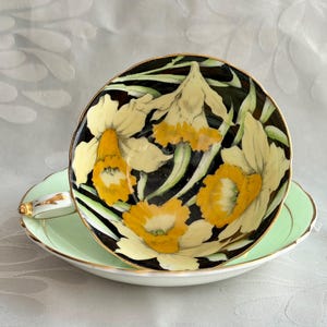 May include: A porcelain teacup and saucer set featuring a black interior decorated with yellow and cream daffodil flowers. The exterior of the cup and the saucer are a pale mint green, and both pieces are finished with a thin gold trim.