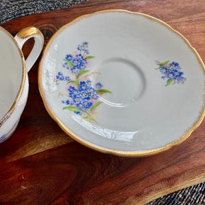 May include: A white teacup and saucer with a gold rim and a floral design featuring blue forget-me-not flowers.