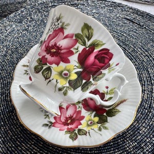 Puede incluir: Una taza de t&eacute; y un platillo blancos con un patr&oacute;n floral. La taza y el platillo presentan un dise&ntilde;o floral rojo, rosa y amarillo con hojas verdes. La taza tiene un borde dorado.