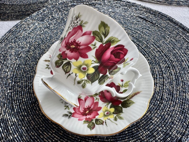 Puede incluir: Una taza de t&eacute; y un platillo blancos con un patr&oacute;n floral. La taza y el platillo presentan un dise&ntilde;o floral rojo, rosa y amarillo con hojas verdes. La taza tiene un borde dorado.