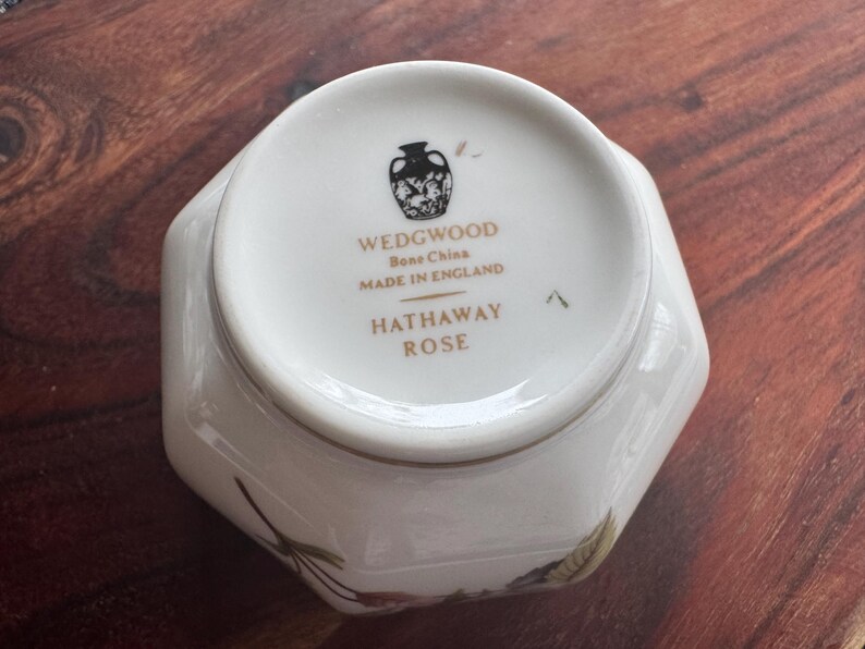 Pode incluir: Um prato de porcelana branca com formato hexagonal. A base do prato apresenta o texto "WEDGWOOD Bone China MADE IN ENGLAND HATHAWAY ROSE" e um emblema decorativo. Detalhes florais s&atilde;o vis&iacute;veis.
