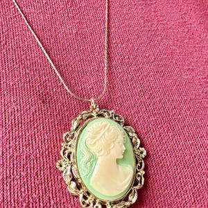 May include: A gold-toned necklace featuring an oval cameo pendant. The cameo has a light green background with a white profile of a woman. The pendant is framed by an ornate gold-colored border.