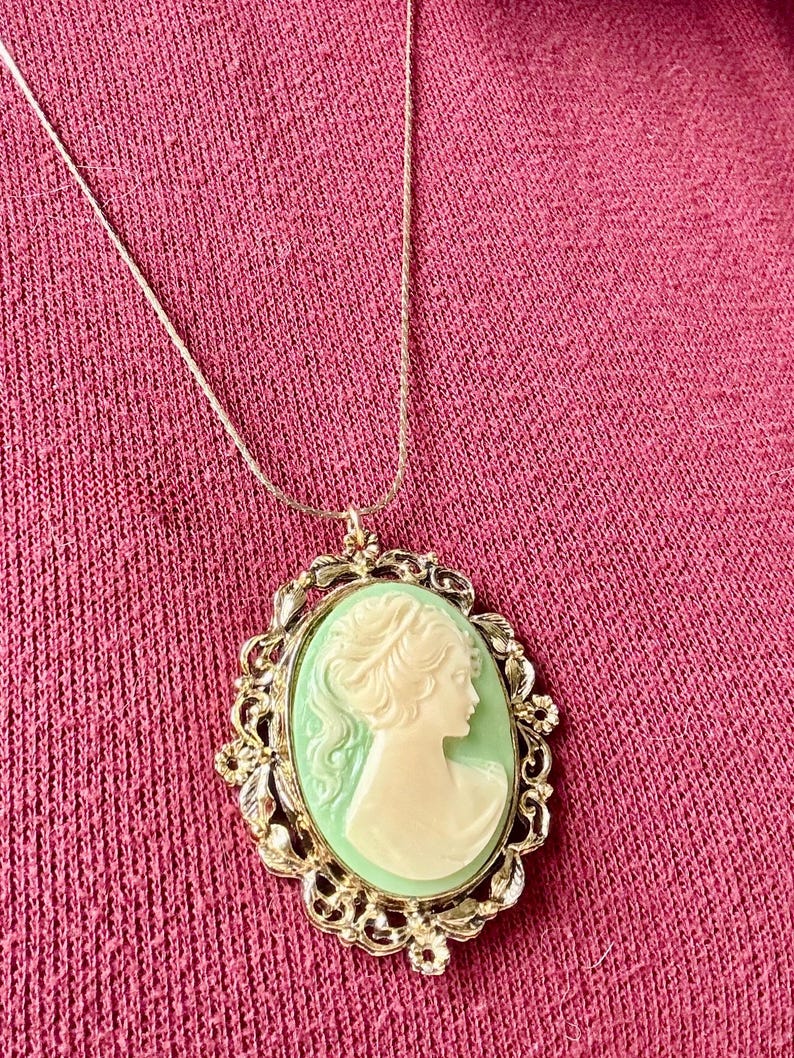 May include: A gold-toned necklace featuring an oval cameo pendant. The cameo has a light green background with a white profile of a woman. The pendant is framed by an ornate gold-colored border.