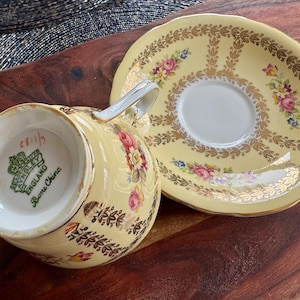 May include: A pale yellow bone china teacup and saucer set featuring a gold leaf pattern and floral motifs. The underside of the teacup displays a green Aynsley England stamp. The set has a scalloped edge and gold trim on the rim and handle.