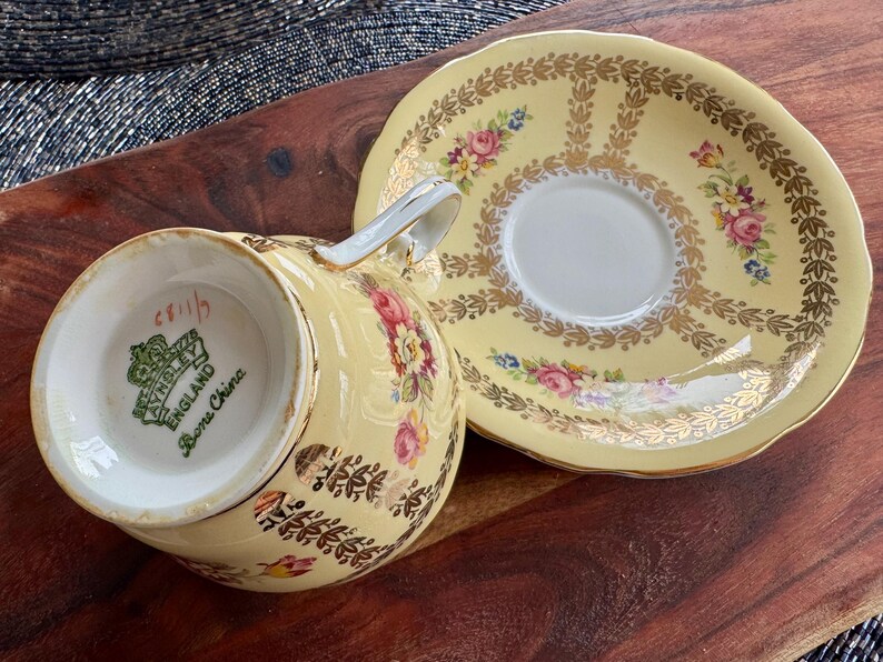 May include: A pale yellow bone china teacup and saucer set featuring a gold leaf pattern and floral motifs. The underside of the teacup displays a green Aynsley England stamp. The set has a scalloped edge and gold trim on the rim and handle.