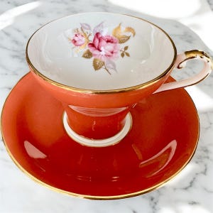 May include: A vintage teacup and saucer set with a floral design. The cup and saucer are a vibrant orange color with a gold trim. The floral design features a pink rose with green leaves.