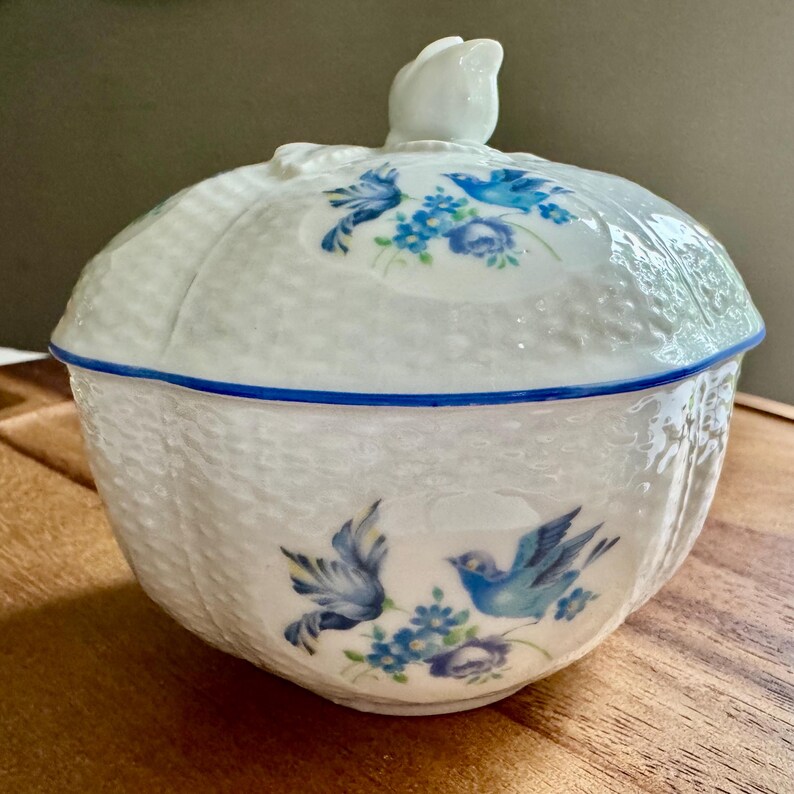 May include: White ceramic lidded bowl with a textured exterior and blue bird and floral designs. The lid features a white tulip-shaped handle and a blue trim. A decorative piece for the home.