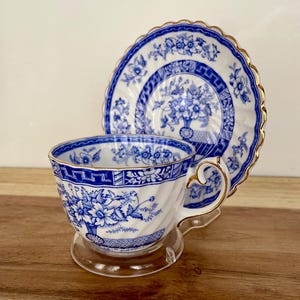 May include: A blue and white porcelain teacup and saucer set. The cup and saucer feature a floral design with gold trim. The teacup is on a clear stand, and the saucer is behind it. The set is likely vintage or antique.
