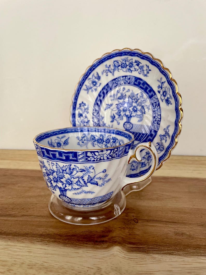 May include: A blue and white porcelain teacup and saucer set. The cup and saucer feature a floral design with gold trim. The teacup is on a clear stand, and the saucer is behind it. The set is likely vintage or antique.