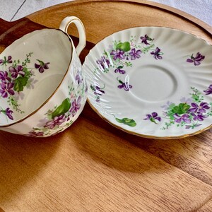 K&ouml;nnte beinhalten: Ein wei&szlig;es Tee-Set mit Tasse und Untertasse mit einem floralen Veilchen-Design. Die Tasse und Untertasse haben einen goldenen Rand. Das Set steht auf einem Holztablett. Die Tasse hat einen Durchmesser von ca. 7.6 cm.