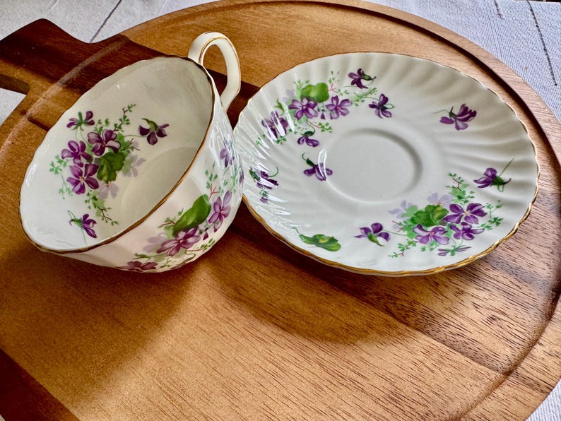K&ouml;nnte beinhalten: Ein wei&szlig;es Tee-Set mit Tasse und Untertasse mit einem floralen Veilchen-Design. Die Tasse und Untertasse haben einen goldenen Rand. Das Set steht auf einem Holztablett. Die Tasse hat einen Durchmesser von ca. 7.6 cm.