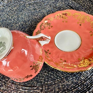 May include: A coral pink teacup and saucer with gold trim and floral designs. The bottom of the teacup has the text "EST. 1775 AYNSLEY FINE BONE CHINA MADE IN ENGLAND" in black.
