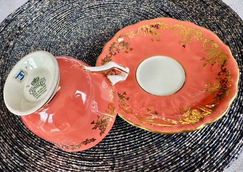 May include: A coral pink teacup and saucer with gold trim and floral designs. The bottom of the teacup has the text "EST. 1775 AYNSLEY FINE BONE CHINA MADE IN ENGLAND" in black.