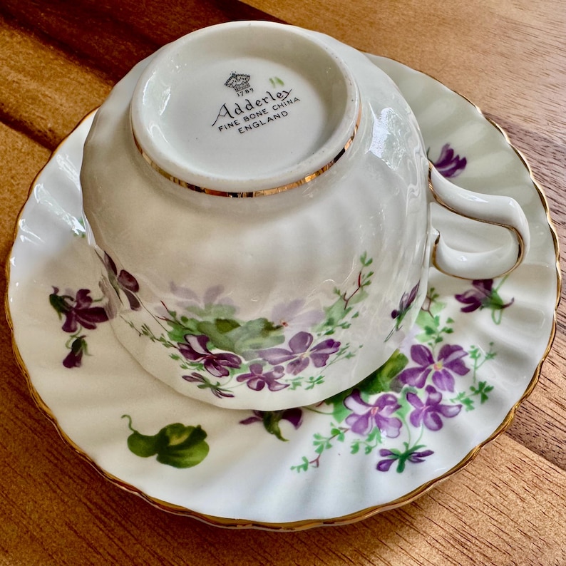 K&ouml;nnte beinhalten: Wei&szlig;es Tee-Set mit floralem Muster aus lila Blumen und gr&uuml;nen Bl&auml;ttern. Die R&auml;nder sind goldfarben verziert. Die Tasse liegt verkehrt herum auf der Untertasse. Hergestellt in England.