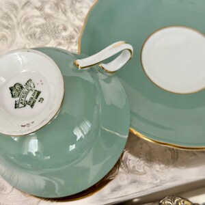 May include: A vintage tea cup and saucer set in a soft teal color with gold trim. The cup has a white interior and a curved handle. The saucer has a white center. The bottom of the cup has the text "AYNSLEY ENGLAND BONE CHINA".