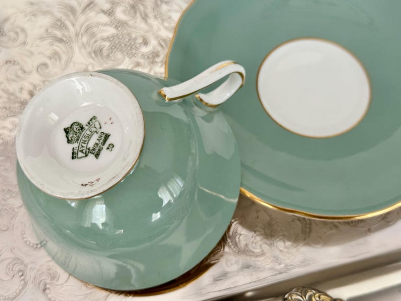 May include: A vintage tea cup and saucer set in a soft teal color with gold trim. The cup has a white interior and a curved handle. The saucer has a white center. The bottom of the cup has the text "AYNSLEY ENGLAND BONE CHINA".