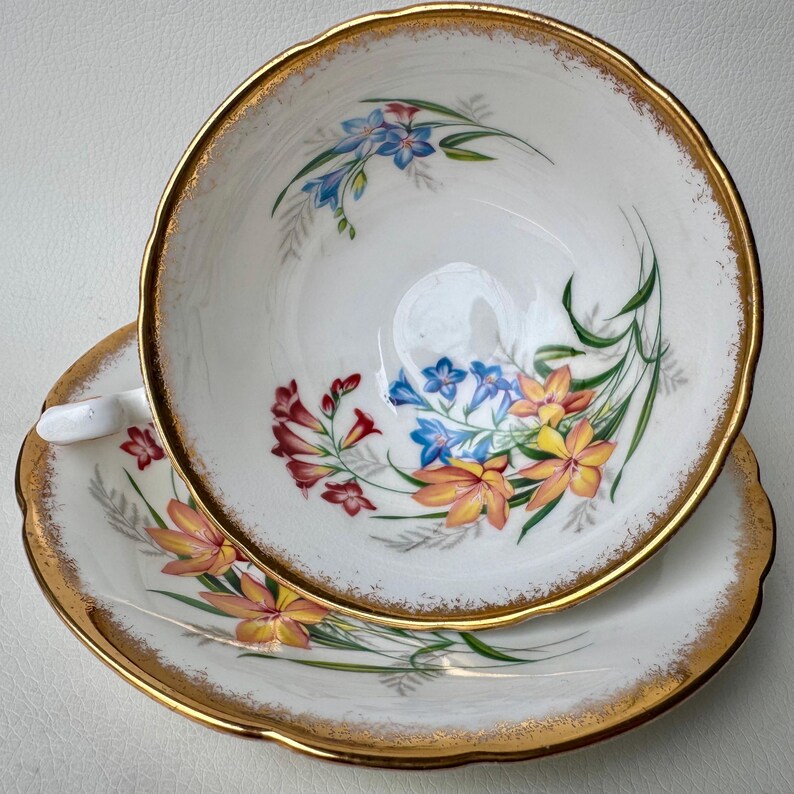 May include: A white teacup and saucer with a gold rim and floral pattern. The flowers are yellow, orange, red, and blue.