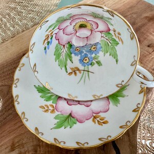 May include: A white teacup and saucer with a floral design. The cup and saucer have a gold trim and feature pink, blue, and green flowers.
