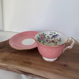 May include: A pink teacup and saucer set. The cup features a floral and bird design inside, with gold trim. The saucer is pink with a white center and gold trim. A classic tea set.