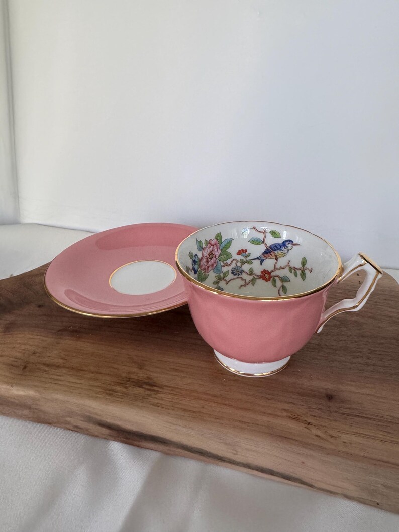 May include: A pink teacup and saucer set. The cup features a floral and bird design inside, with gold trim. The saucer is pink with a white center and gold trim. A classic tea set.