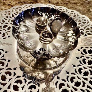 May include: A silver-toned, tiered serving tray with ornate, openwork detailing. Three rings with various designs rest in the top bowl. The tray is likely used for displaying jewelry or small items.