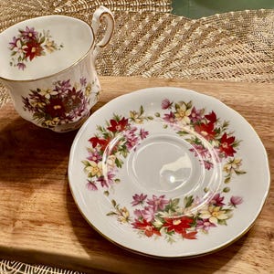 May include: A white teacup and saucer set with a floral pattern in shades of pink, red, yellow, and green. The floral design features a variety of flowers, including roses, daisies, and other wildflowers.
