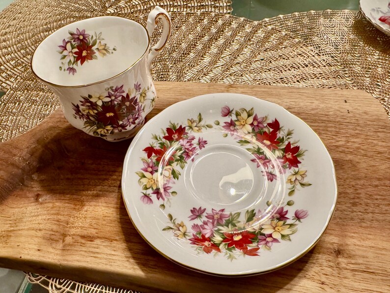 May include: A white teacup and saucer set with a floral pattern in shades of pink, red, yellow, and green. The floral design features a variety of flowers, including roses, daisies, and other wildflowers.