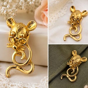 May include: A gold-toned mouse brooch with round glasses and a curled tail. The mouse has detailed features and is posed upright. The brooch is shown on a white surface, a green fabric, and a white lace.