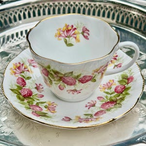 May include: White teacup and saucer set with a floral design featuring pink and yellow flowers and green leaves. The cup and saucer have gold trim. The set is placed on a silver tray.