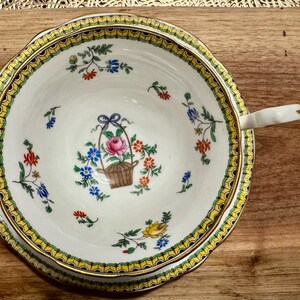 Pu&ograve; includere: Una tazza da t&egrave; e un piattino bianchi con un bordo dorato e un motivo floreale. Il motivo floreale presenta fiori rosa, gialli e blu con foglie verdi. Il piattino ha un bordo ondulato.