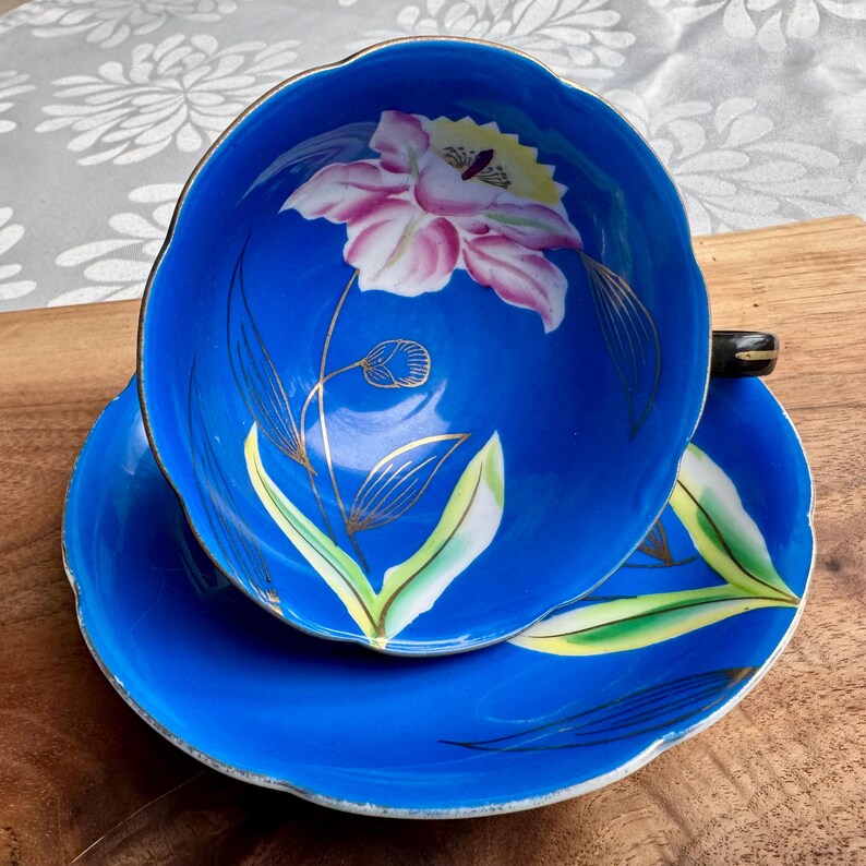 Puede incluir: Un juego de taza y platillo de t&eacute; azul con un dise&ntilde;o floral. La taza y el platillo est&aacute;n decorados con una flor rosa con detalles dorados sobre un fondo azul.