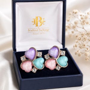 Op de afbeelding: Hartvormige oorbellen in een marineblauwe doos. De oorbellen hebben een goudkleurige setting met paarse, roze en turquoise hartvormige stenen, geaccentueerd met heldere kristallen. De doos is van Bradford Exchange.