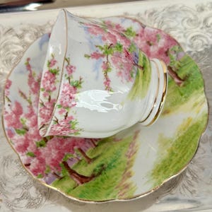 May include: A white teacup and saucer set with a floral design. The cup and saucer feature pink cherry blossoms, green grass, and gold trim. The teacup is resting on the saucer, showcasing the detailed artwork.